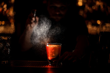 Professional bartender spraying to the cocktail glass with one big ice cube in the dark