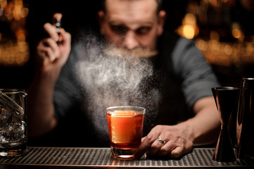 Bartender spraying to the cocktail glass with one big ice cube under the light