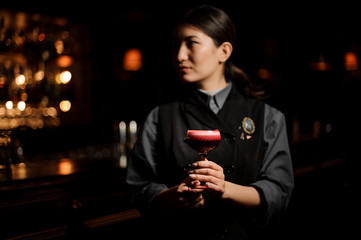Professional female bartender serving a smooth crimson cocktail in the glass with a one pink rose bud as a decor