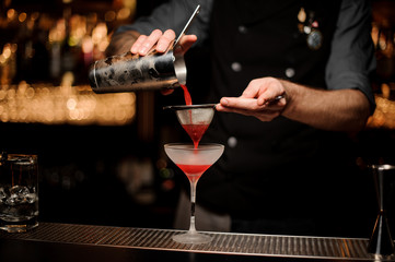 Professional bartender pouring a smooth crimson cocktail through the sieve to the glass