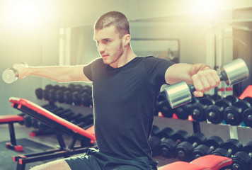 Athletic young man exercising his shoulders and doing side raises with pair of dumbbells at gym