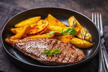 Grilled beef steak with potatoes and basil in a black plate on dark background.