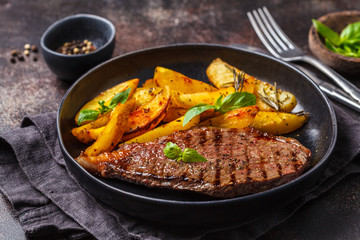 Grilled beef steak with potatoes and basil in a black plate on dark background.
