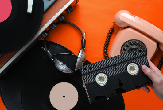 Female hand holds video cassette. 80s style. Vinyl player, headphones, rotary phone on orange  background. Top view, flat lay