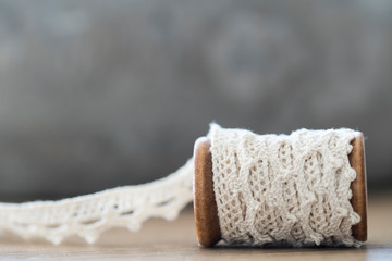 close up wooden spool with white lace, ribbon, on wooden table, grey background. Space for text
