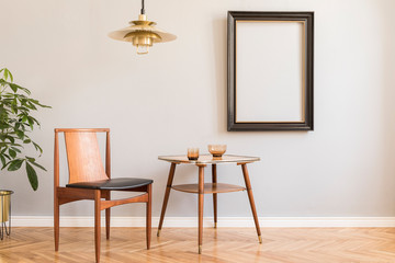 Stylish and design retro interior with design chair, gold lamp, small table with accessories. Black mock up frame on the grey background wall. Minimalistic concept of sitting room. Real photo. 