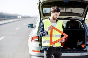 Man in road vest holding emergency triangle sign near the broken car on the roadside