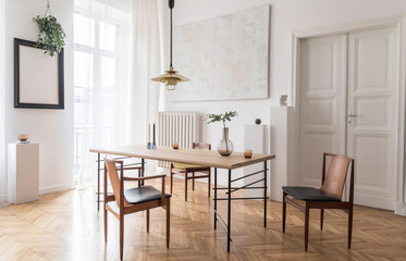 Fototapeta na wymiar Stylish and modern dining room interior with design sharing table, chairs, gold pendant lamp, abstract paintings and elegant accessories. Tropical leafs in vase. Eclectic decor. Brown wooden parquet.