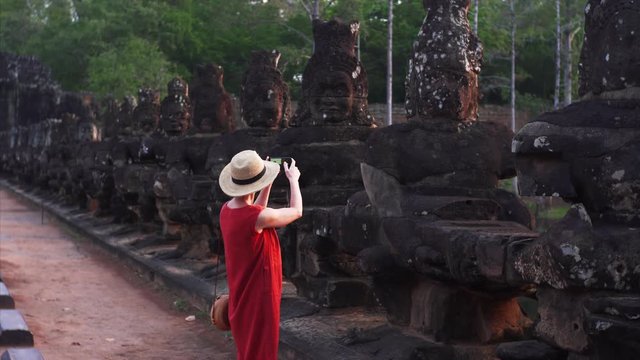 Attractive young woman walking through the temple and taking pictures morning. Ancient statues on South Gate bridge of Angkor Thom temple.
