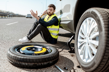 Man calling the road assistance sitting near the car with broken wheel on the roadside