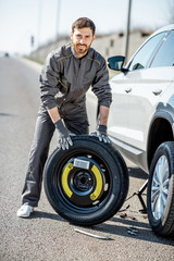 Portrait of a handsome road assistance worker in uniform standing with spare wheel near the broken...