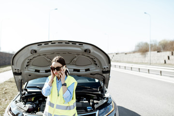 Embarrassed woman calling road assistance standing near the broken car on the roadside of the highway