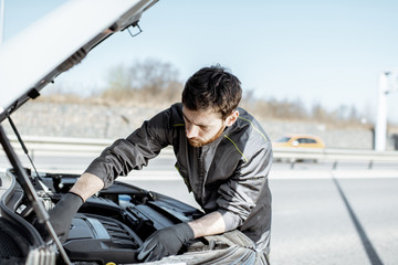 Handsome auto mechanic in uniform repairing engine of the broken car on the road