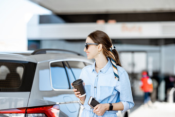 Young woman having a coffee break standing near the car on the gas station