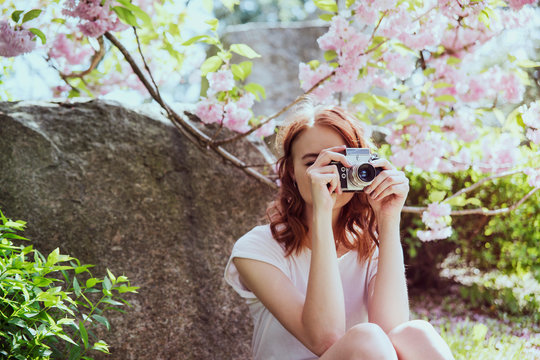 Young beautiful redhead girl in white t-shirt and gray skirt takes a photo in springtime blossom cherry trees garden. Hobby, vlogger, blogger, analog photo, social media.  Spring time for a walk.