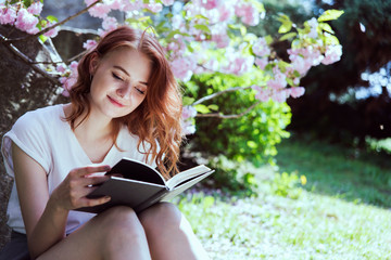 Young beautiful redhead girl in white t-shirt and gray skirt reads a book for her blogg channel in...