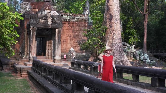 Attractive young woman walking through the temple morning. Woman in the red dress strolling through the temple in Cambodia, Angkor, Ta Prohm Temple. 