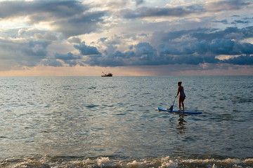 Young female paddling on SUP board in the sea at amazing dark sunset and waves. Sup surfing woman silhouette - active recreation in nature. 