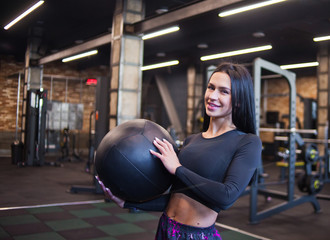 Cheerful brunette woman in sportswear holding medicine ball in her hand and posing at camera .