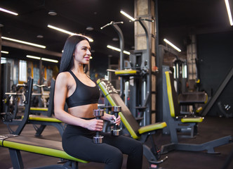 Fototapeta na wymiar Smilling young woman holds dumbbells in hands while sitting on a bench in gym
