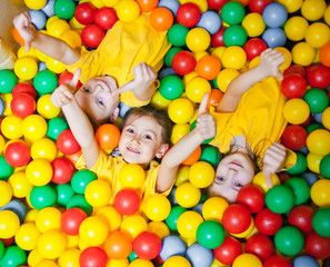 Three happy little kids girls in ball pit smiling happily at camera and show thumb up while having fun in children play center. Top view
