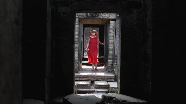 Attractive young woman walking through the coridors of the temple and taking pictures morning. Woman in the red dress taking pictures of the temple in Cambodia, Angkor, Ta Prohm Temple. 