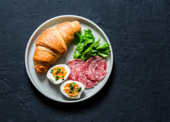 Delicious brunch or breakfast - croissant, boiled egg, salami sausage, green salad on a dark background, top view