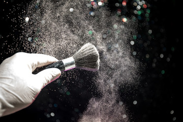 Makeup brush in hand with cosmetic powder on dark background with light and smoke. Powder splash on dark