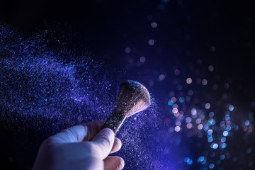 Makeup brush in hand with cosmetic powder on dark background with light and smoke. Powder splash on dark