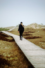 Back view silhouette of a young girl walking on a wooden path with fresh green nature dunes landscape in a moody fog weather. National Park Darß, Zingst, Baltic Sea (Deutsche Ostsee) in Germany