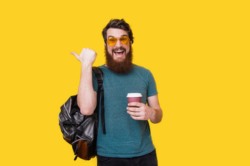 Bearded man wearing stylish glasses, with backpack pointing away, holding cup of coffee