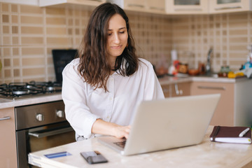 Smiling young woman using laptop in the kitchen at home, freelancer concept