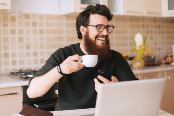 Young man with beard typing on laptop. He is sitting in the kitchen and smiling because of seen video. Happy boy is trying to drink some coffee