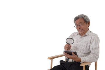 An Asian old man sitting on a chair and using a magnifying glass to look at a tablet with eye sight problem on isolated white background.
