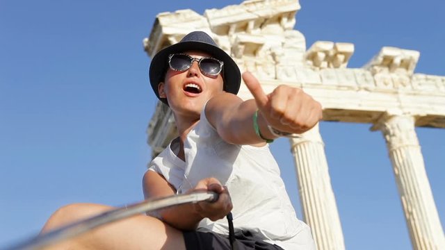 Girl on vacation taking selfie picture of herself and showing thumbs up gesture. Background of ancient ruins.
