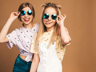 Two young beautiful smiling blond hipster girls in trendy summer colorful T-shirt clothes. Sexy carefree women posing near beige wall in round sunglasses. Positive models showing tongue