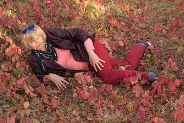 A happy middle-aged woman is in the forest in early autumn. Pink clothes.Lies on the grass. There are large pink leaves around.