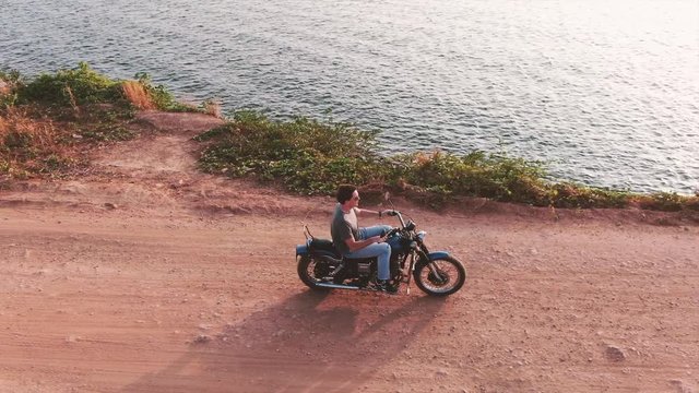 Aerial profile view follow motorcycle rider along a dirt road by a river at sunset during a travel adventure.