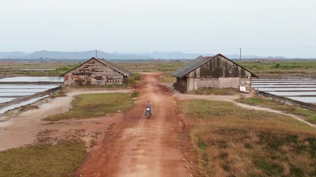 Wide aerial establishing shot offroad motorcycle rider at sunset rides down a dirt road in Asia. Salt fields can be seen nearby, with large wooden sheds.