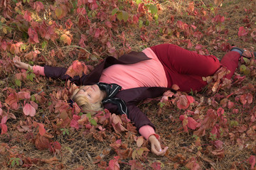 A happy middle-aged woman is in the forest in early autumn. Pink clothes.Lies on the grass.There are large pink leaves around.