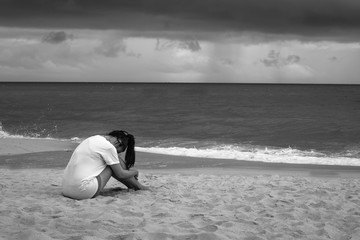 Young sad woman sitting on the beach crying in the rain. Black and white