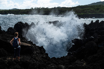 Woman hiker stands on the volcanic sharp ground and watches giant splash of the water caused by...