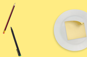 Blank square paper sticker with curved corner in white round plate near wooden pencil and black pen on yellow table. Top view. Menu template, business or education concept. Copy space