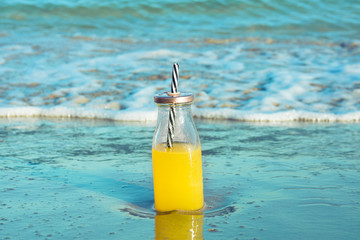 Glass bottle with fresh tropical fruits juice with straw standing on beach sand shore blue turquoise foamy sea waves. Golden sunlight. Summer vacation relaxation wanderlust