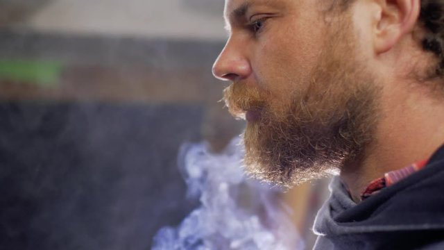 a worker with a beard and mustache smokes a cigarette indoors against a backlight. 4 k
