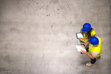 Top view of two industrial workers wearing hardhats and reflective jackets holding tablet and...