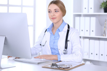 Fototapeta na wymiar Young woman doctor at work in hospital looking at desktop pc monitor. Physician controls medication history records and exam results. Medicine and healthcare concept