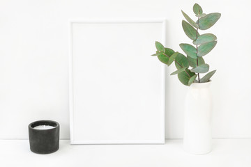 White frame mockup composition with branches of green silver dollar eucalyptus in ceramic vase black candle on table wall background. Copy space for text lettering artwork. Elegant minimalist