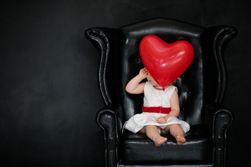 Little blonde girl sitting on the armchair with red heart balloon on the St. Valentine's day