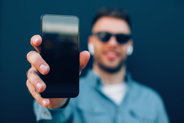 Young man listening to music via wireless earphones and smartphone leaning against a blue wall, seledtive focus on smartphone